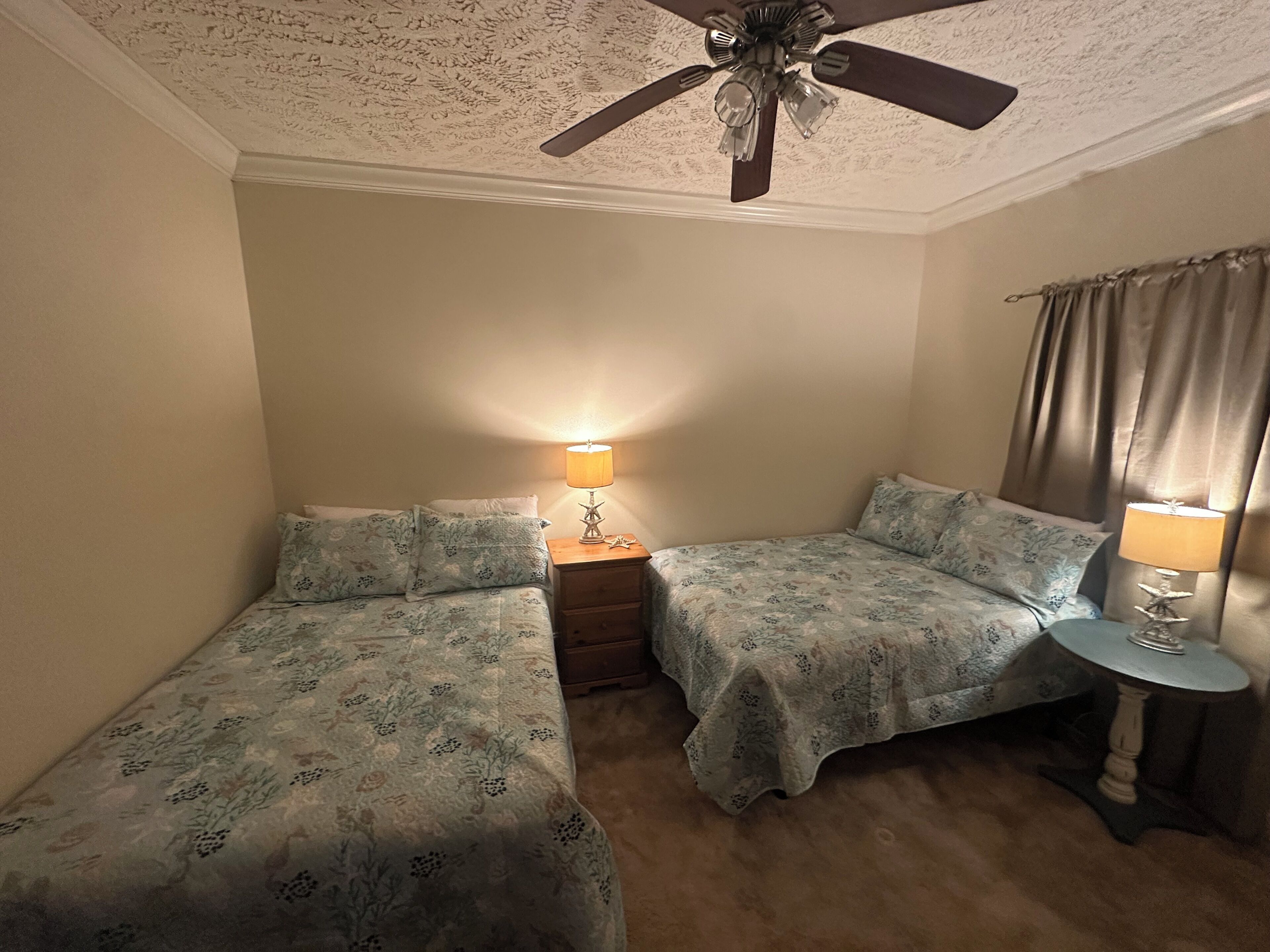 A bedroom with two beds, a ceiling fan, a nightstand with a lamp, and a window with curtains.