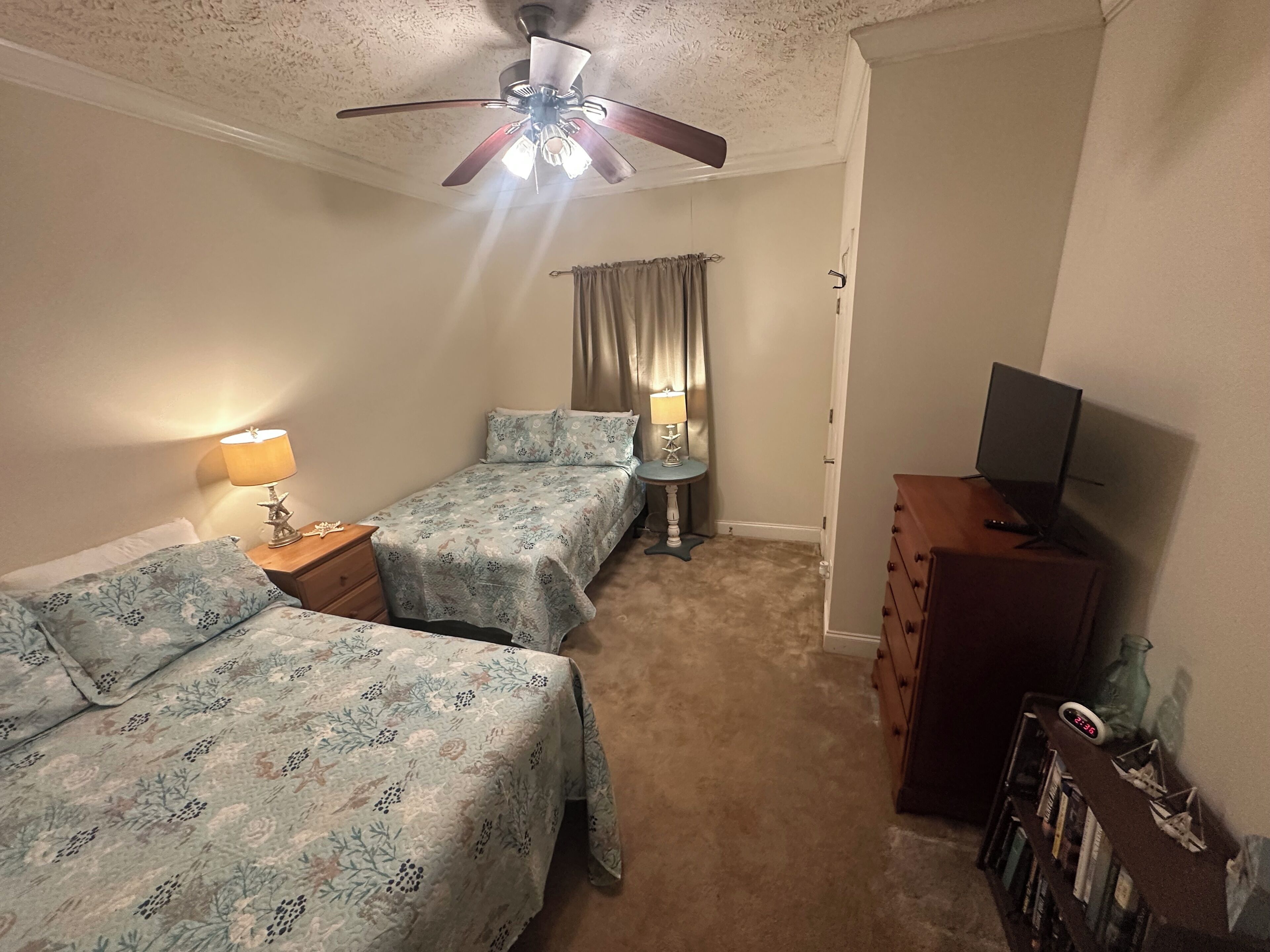 Ein Schlafzimmer mit zwei Betten, einem Deckenventilator, einem Schreibtisch mit Lampe, einem Schminktisch und einem Bücherregal.