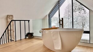 Een modern interieur met een groot bad, een houten trap en zicht op besneeuwde bomen door grote ramen.