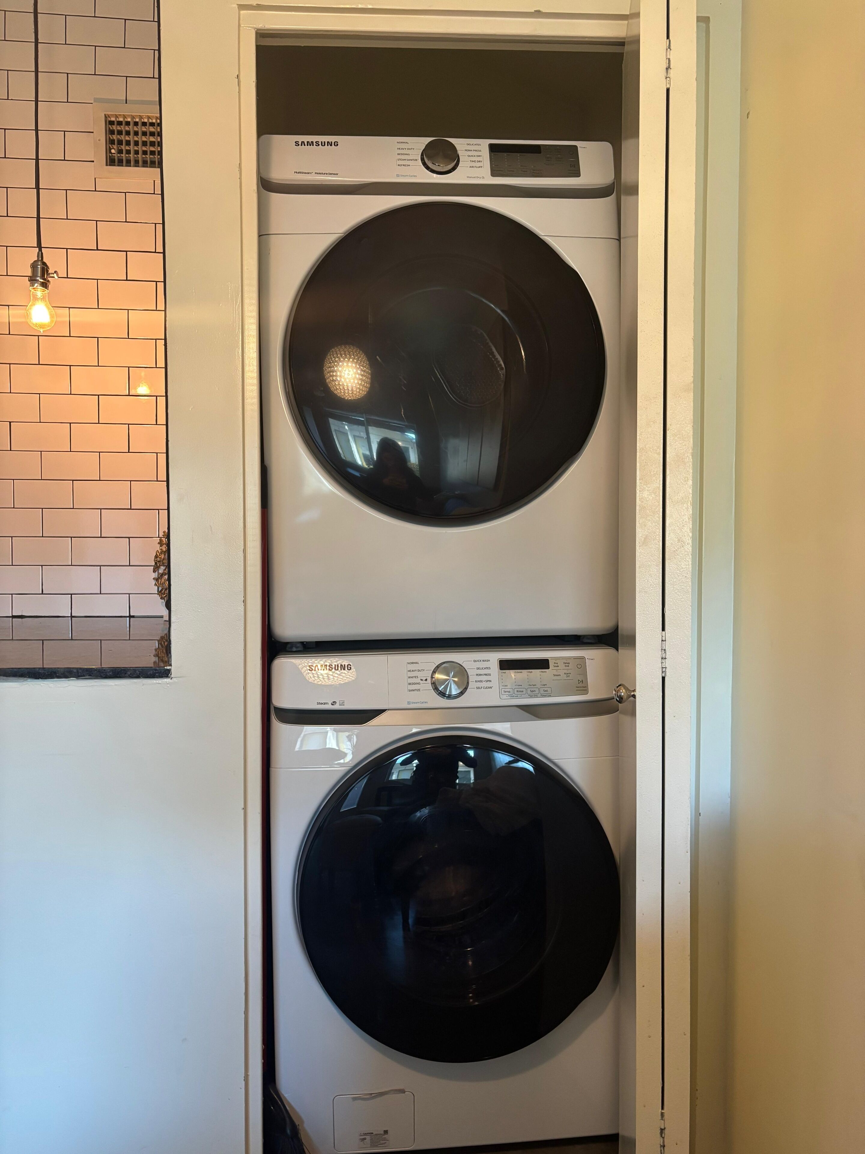 Un appareil combiné lave-linge et sèche-linge à chargement frontal de la marque Samsung, avec un mur décoré d’un motif imitant des briques et un luminaire suspendu.