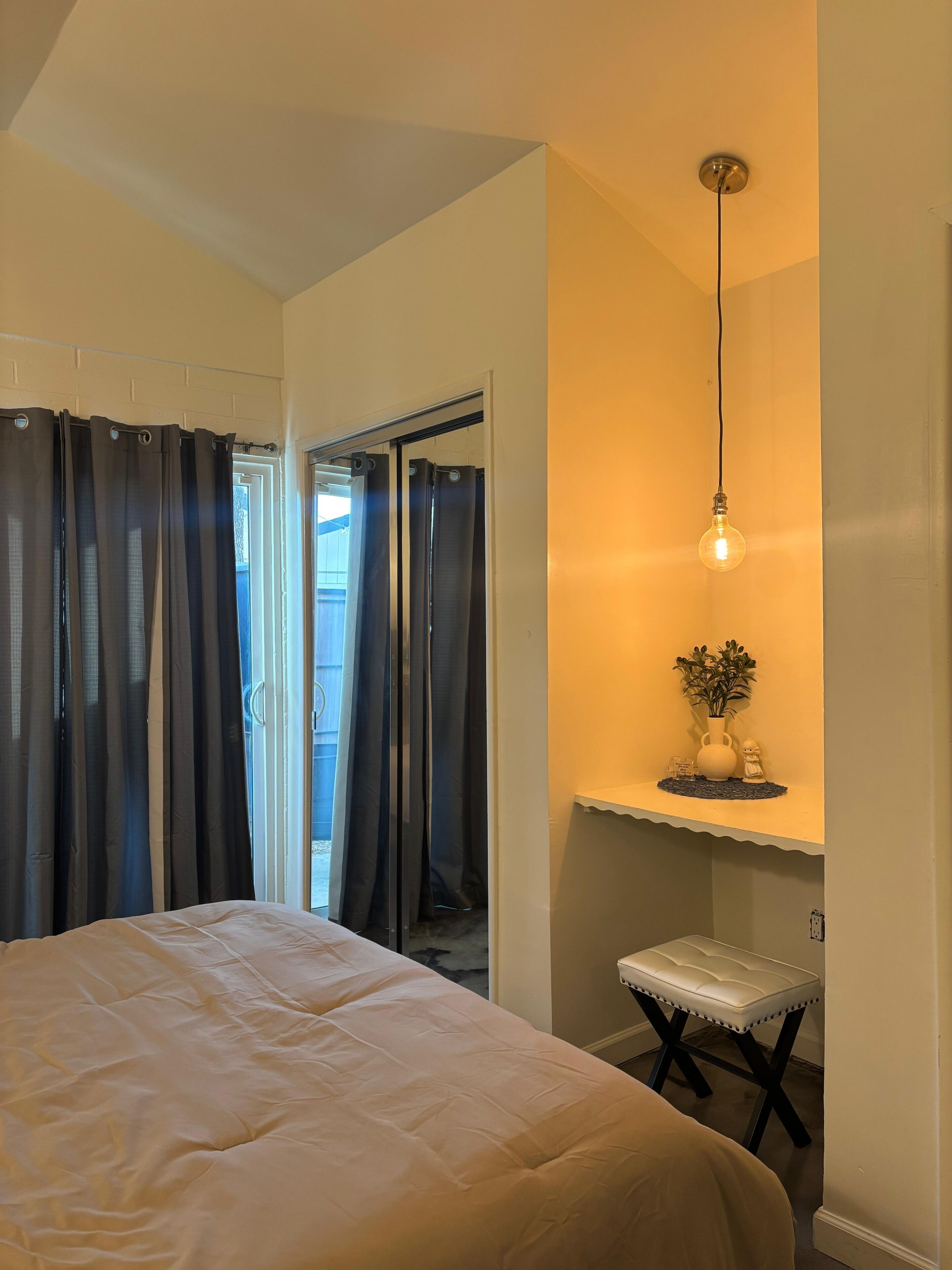 A bedroom with a bed, a desk with a plant, a hanging light, and a sliding door.