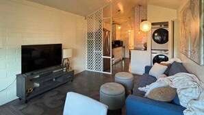 A modern living room with a flat-screen TV, a sofa, ottomans, and a dining area with a wooden table.