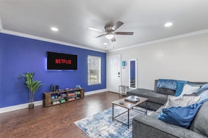 A living room with a flat-screen TV, a ceiling fan, a bookshelf, a gray sofa, and a blue accent wall. - Stadium View Balcony | Walk to AT&T + Globe Life! (Arlington)