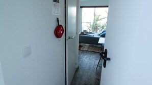 Un pasillo con una puerta blanca, un extintor rojo y una ventana que da a un balcón con vistas a palmeras.