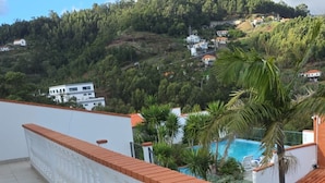 Varanda com guarda-corpo branco e tijolos vermelhos, com vista para uma piscina e uma colina com casas.