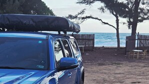 Un SUV bleu est garé sur un chemin de terre, avec une planche de surf sur le toit, à proximité d’une clôture en bois et de chaises de plage.