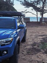 A blue SUV parked on a dirt path with a surfboard on the roof, near a wooden fence and beach chairs.