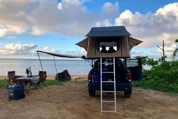 Una tienda de campaña con una escalera, un vehículo azul y una mesa con sillas en una playa arenosa.