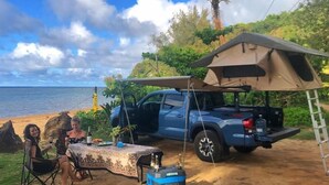 Un coin de camping aménagé sur la plage, avec une tente, une table avec des boissons et un camion bleu.