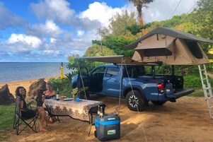 A beachside camping setup with a tent, a table with drinks, and a blue pickup truck.