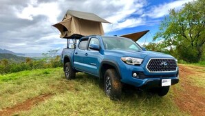 Un camion bleu avec une tente fixée à l’arrière, garé sur une pelouse, avec un ciel dégagé et des collines lointaines en arrière-plan.