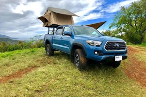 A blue pickup truck with a tent attached to the back, parked on a grassy area with a clear sky and distant hills in the background.
