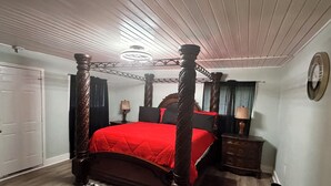 A four-poster bed with a red bedspread, a wooden floor, a patterned rug, a nightstand with a lamp, and a wall clock.