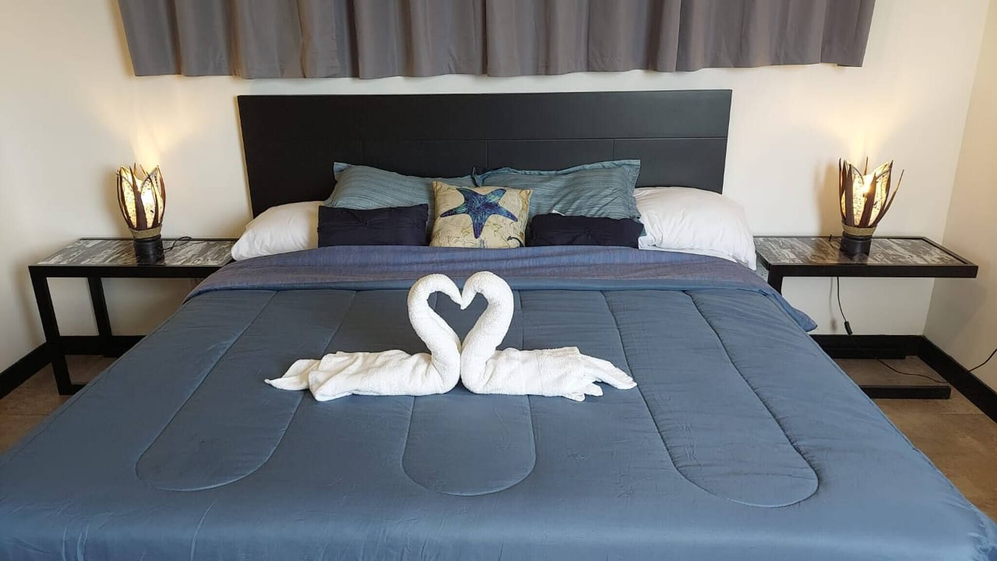 Un lit bien fait, agrémenté d’une décoration en forme de cœur réalisée avec des serviettes, une tête de lit sombre et deux tables de chevet avec des lampes.