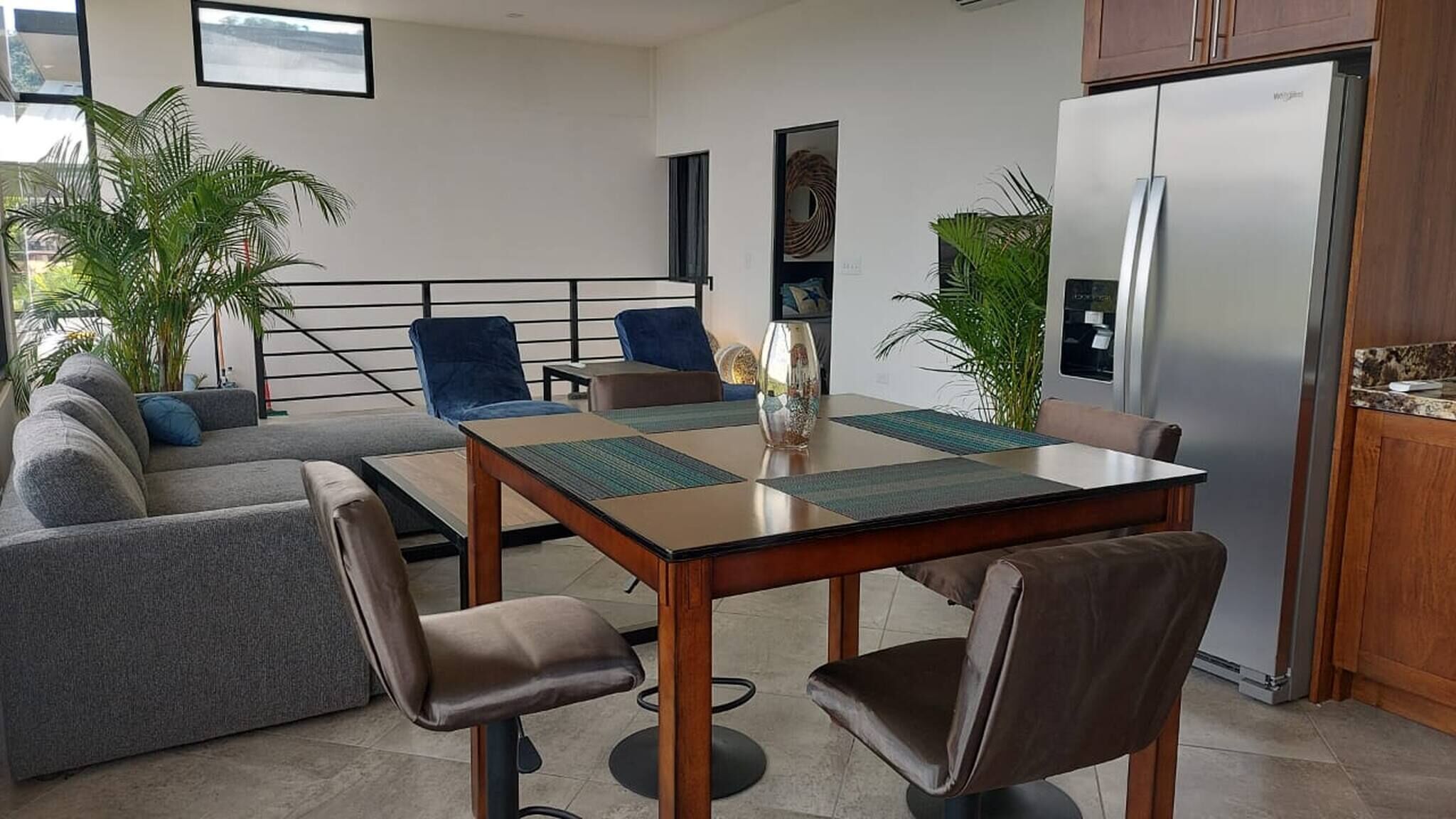 Et moderne køkken med et køleskab, spisebord og stole. Der er planter og en sofa i opholdsstuen.