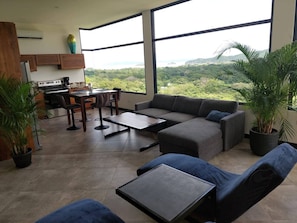 A modern living room with a sofa, coffee table, and dining area, large windows with a view of greenery and hills. - Modern, Deluxe, Ocean View Guest House With Pool (Sardinal)