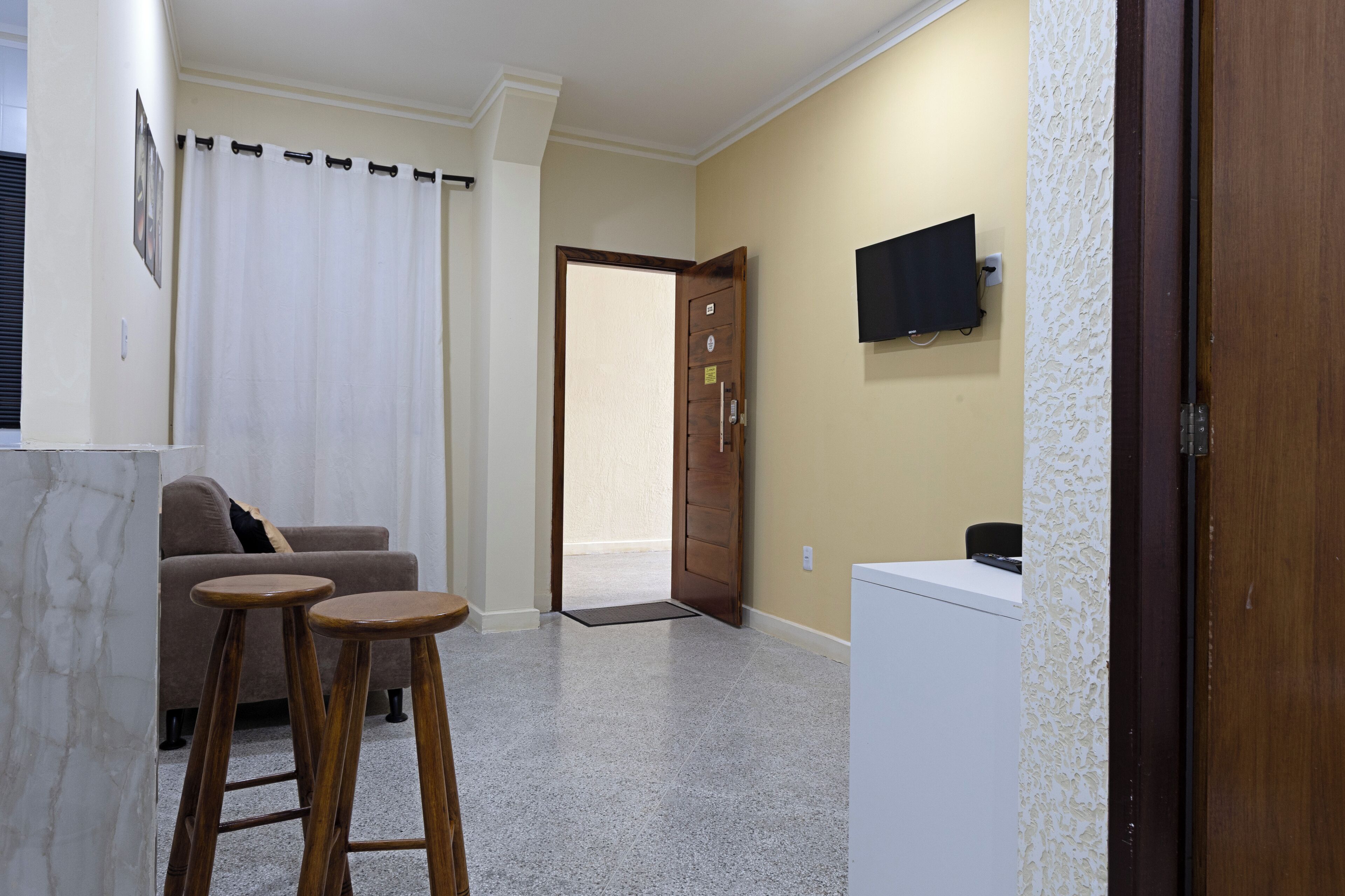 A room with a sofa, two stools, a television on the wall, a door, and a small white appliance.