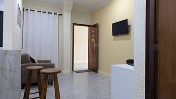 A room with a sofa, two stools, a television on the wall, a door, and a small white appliance.