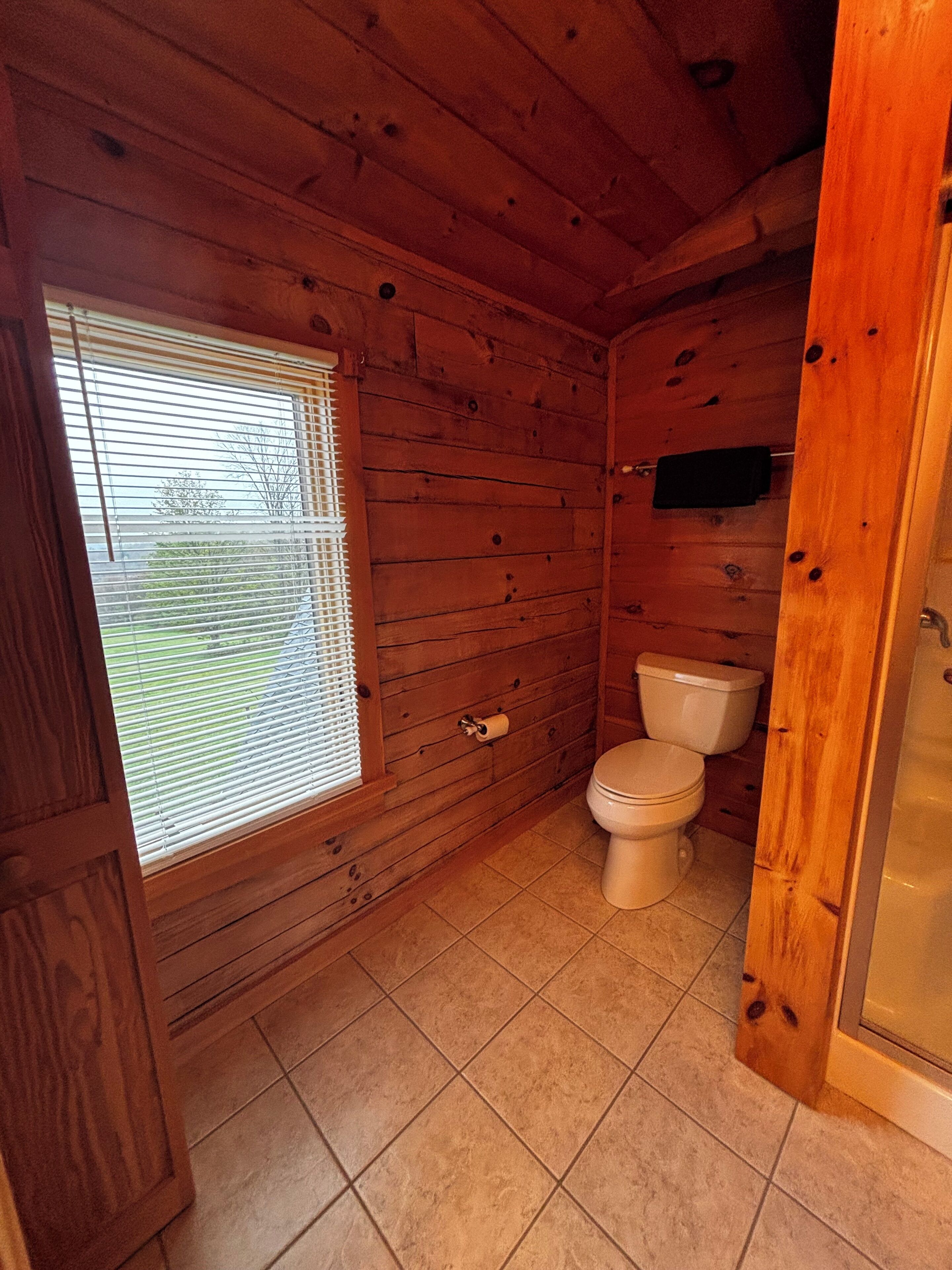 A wooden cabin bathroom with a toilet, a window with blinds, and a shower area.