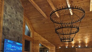 Un vaste espace de vie agrémenté d’une cheminée en pierre, de poutres apparentes en bois, d’un grand lustre et d’un téléviseur diffusant une chaîne d’information.