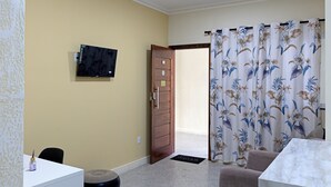 Ein Zimmer mit an der Wand montiertem Fernseher, einer Holztür, einem blumigen Vorhang, einem weißen Tisch und einem schwarzen Stuhl.