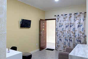 A room with a TV mounted on the wall, a wooden door, a floral curtain, a white table, and a black chair. - Laize & Lucas Residential - 102 - Comfort Minutes from the Beach (Aracaju)