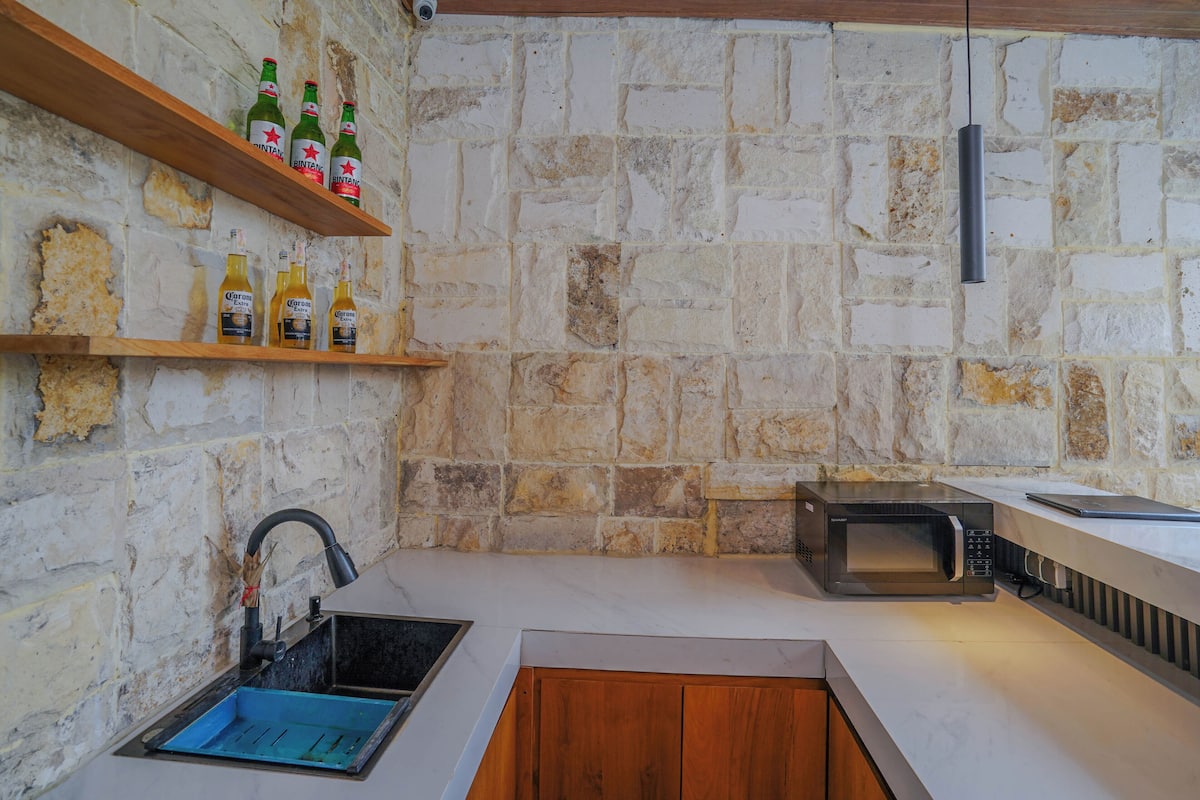 Una cocina con pared de piedra, un lavamanos, un microondas y repisas con botellas.