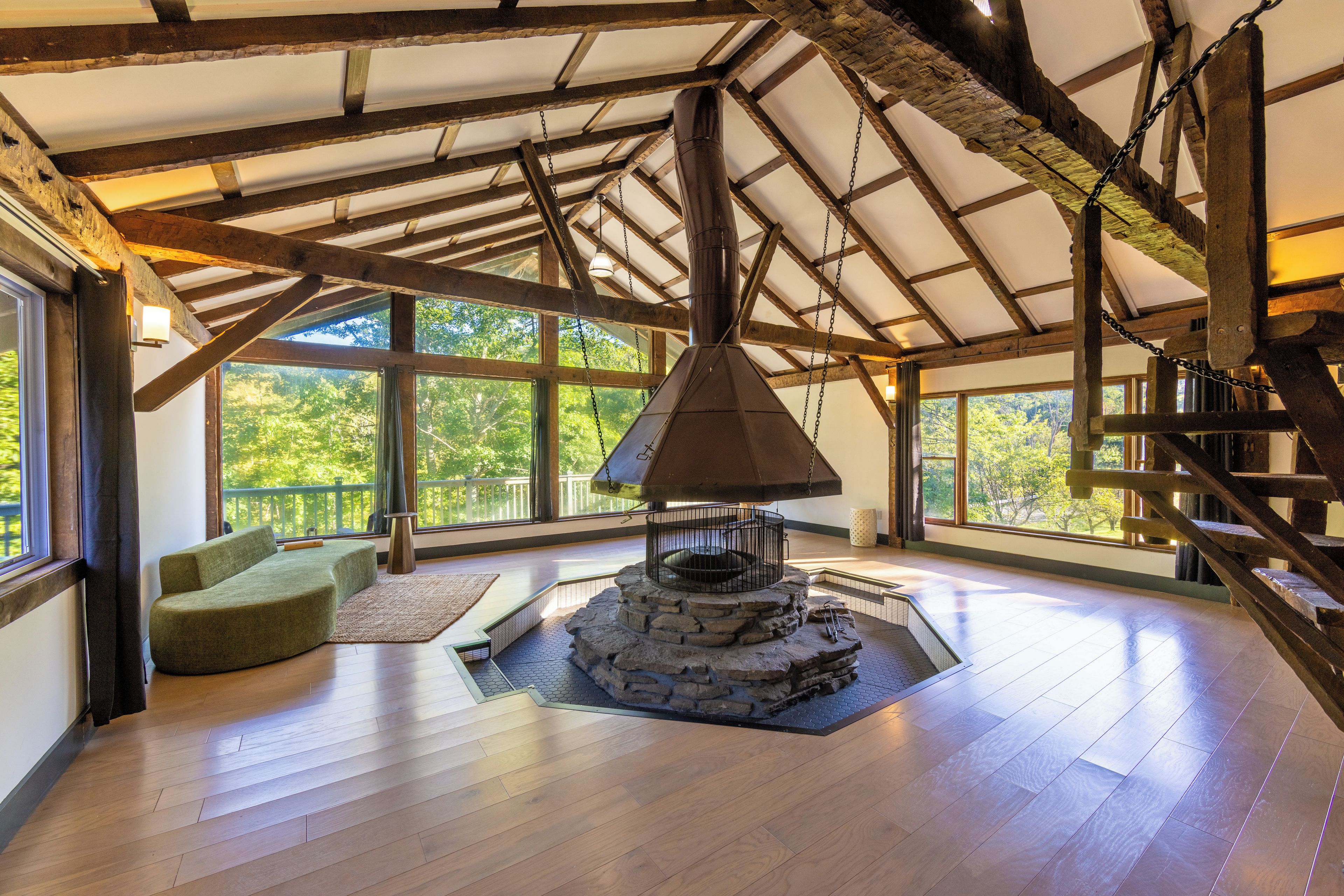 A spacious room with a stone fireplace, wooden beams, and large windows offering a view of trees.
