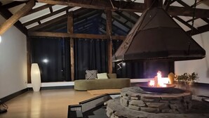 A room with a stone fire pit, a sofa, a large window, and wooden beams. - Beaverkill Riverfront| Fly Fishing, Golf & Firepit (Roscoe)