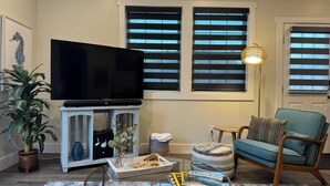 A living room with a flat-screen TV, a blue armchair, a glass coffee table, a potted plant, and a lamp.