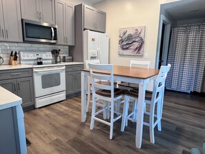 A modern kitchen with grey cabinets, a microwave, a stove, and a refrigerator. There is a wooden dining table with white chairs.