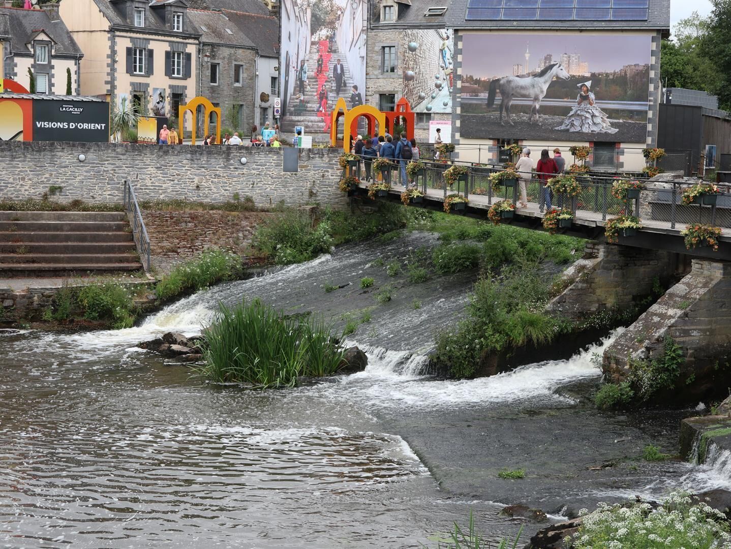A river flowing through a town with stone steps and a pedestrian bridge adorned with flowers.