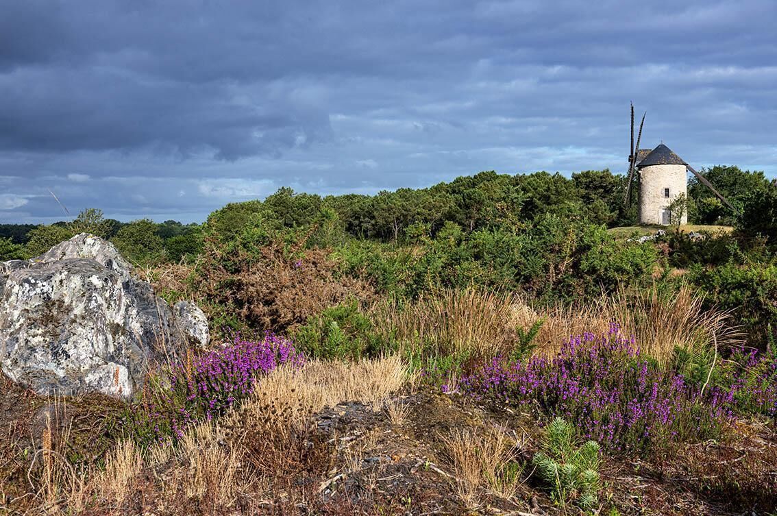 A stone windmill on a hill with surrounding vegetation and a large rock.