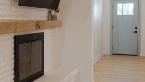A room with a fireplace, a mounted TV, and a door leading to another room. - Bagby Bunkhouse • Beautiful Cottage 7 minutes from Baylor and Magnolia • Pond (Waco)