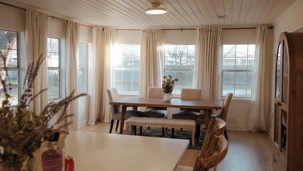 A dining room with a wooden table, chairs, and a bench. There are large windows with curtains, a vase with flowers, and a wooden cabinet. - Bagby Bunkhouse • Beautiful Cottage 7 minutes from Baylor and Magnolia • Pond (Waco)