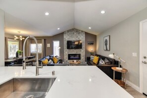 Une cuisine moderne avec un îlot central, un coin repas avec une table et des chaises, un salon avec un canapé et une cheminée, et un téléviseur fixé au mur.