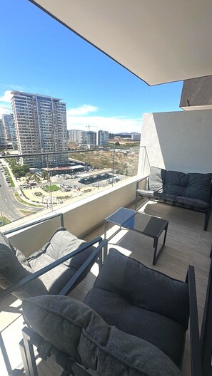 Ein Balkon mit einer Couch und Stühlen, einem Tisch sowie Blick auf eine Stadtlandschaft mit Gebäuden und einer Straße.