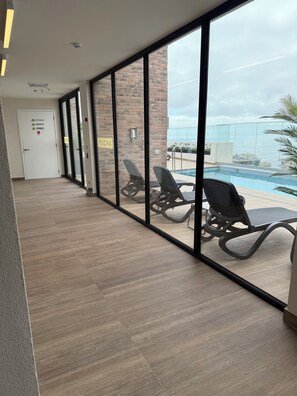 A corridor with glass walls leading to a pool area, featuring lounge chairs and a clear view of the pool. - Nuevo Apartamento cerca de la playa en Reñaca con parking y piscina. (Viña del mMar)