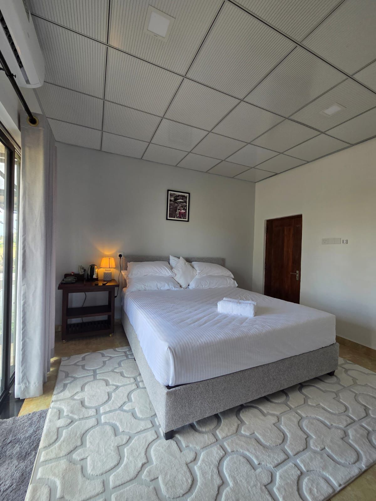 A hotel room with a large bed, a bedside table with a lamp, a framed picture on the wall, and a door.