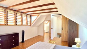 A bedroom with a sloped ceiling, wooden beams, a bed, a dresser, and a window with blinds.