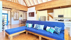 A spacious living area with a wooden bench, a blue sofa with patterned cushions, a kitchen with white cabinets, and a dining area with wooden chairs and a table.