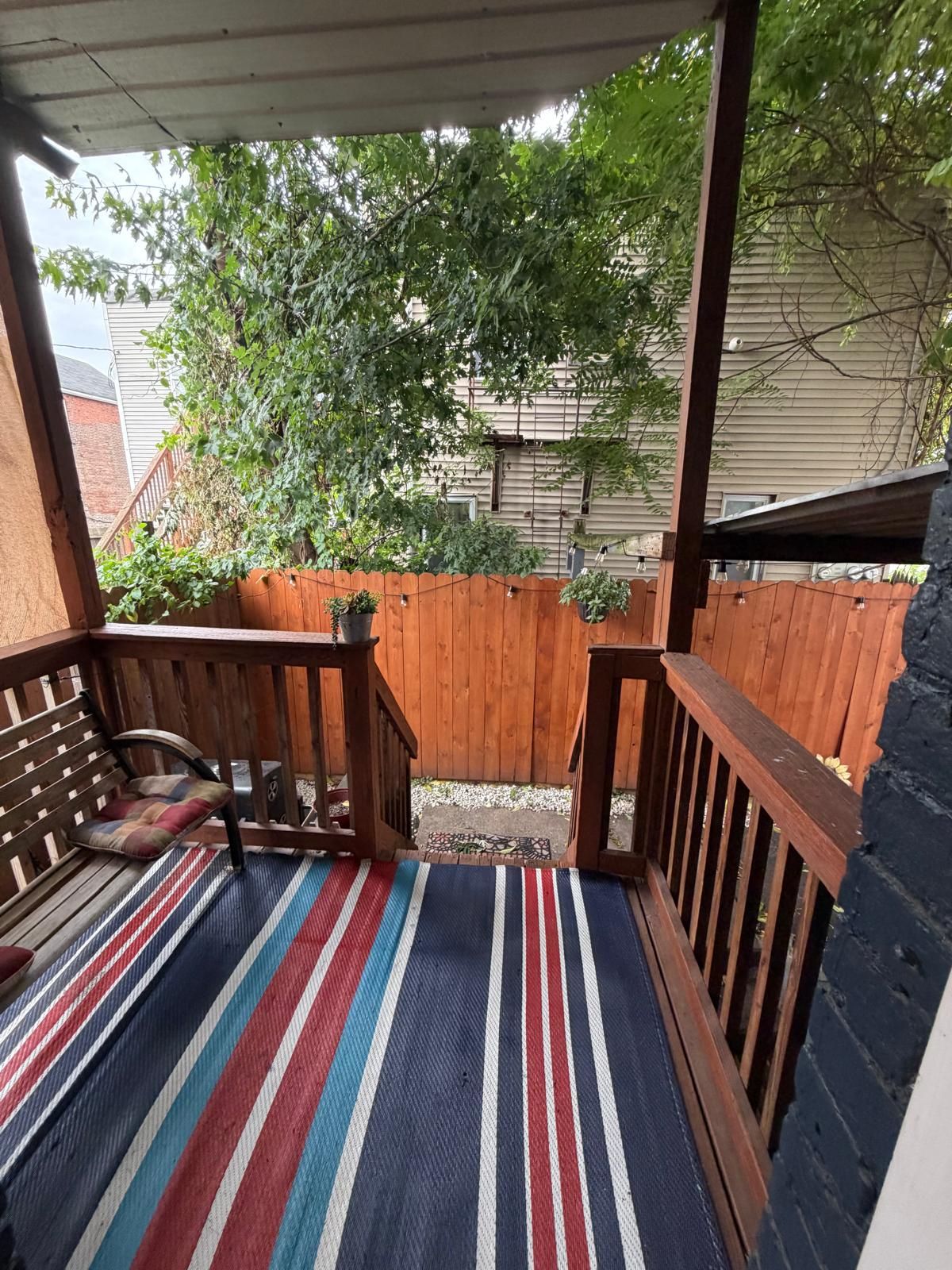 Un espacio exterior techado con una terraza de madera, un banco y un colorido tapete a rayas.