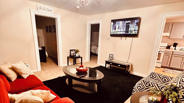 Una sala de estar con un sofá rojo, una mesa de centro, un televisor montado en la pared y una cocina visible a través de una puerta abierta.
