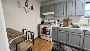 Una cocina con armarios grises, una estufa blanca, un fregadero y una zona de comedor con una silla y una mesa.