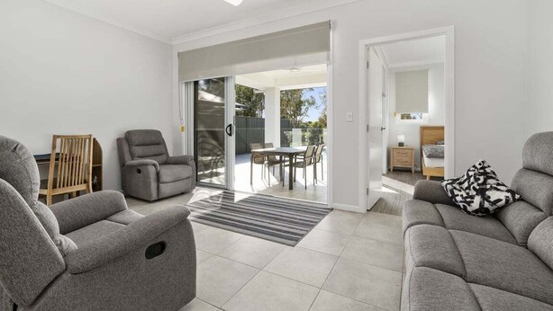 Un salon moderne avec des canapés gris, un fauteuil en bois, un coin repas comprenant une table et des chaises, et une porte-fenêtre coulissante donnant sur une terrasse extérieure.