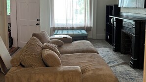Un salon avec un canapé d’angle beige, un ventilateur de plafond, une fenêtre avec des rideaux légers, un tél éviseur posé sur un meuble et une cheminée.