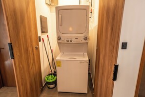 A compact, white washing machine in a narrow hallway with wooden doors and a mop leaning against the wall. - Cozy retreat in Mazatlan | Close to beach, restaurants and boardwalk (Mazatlán)