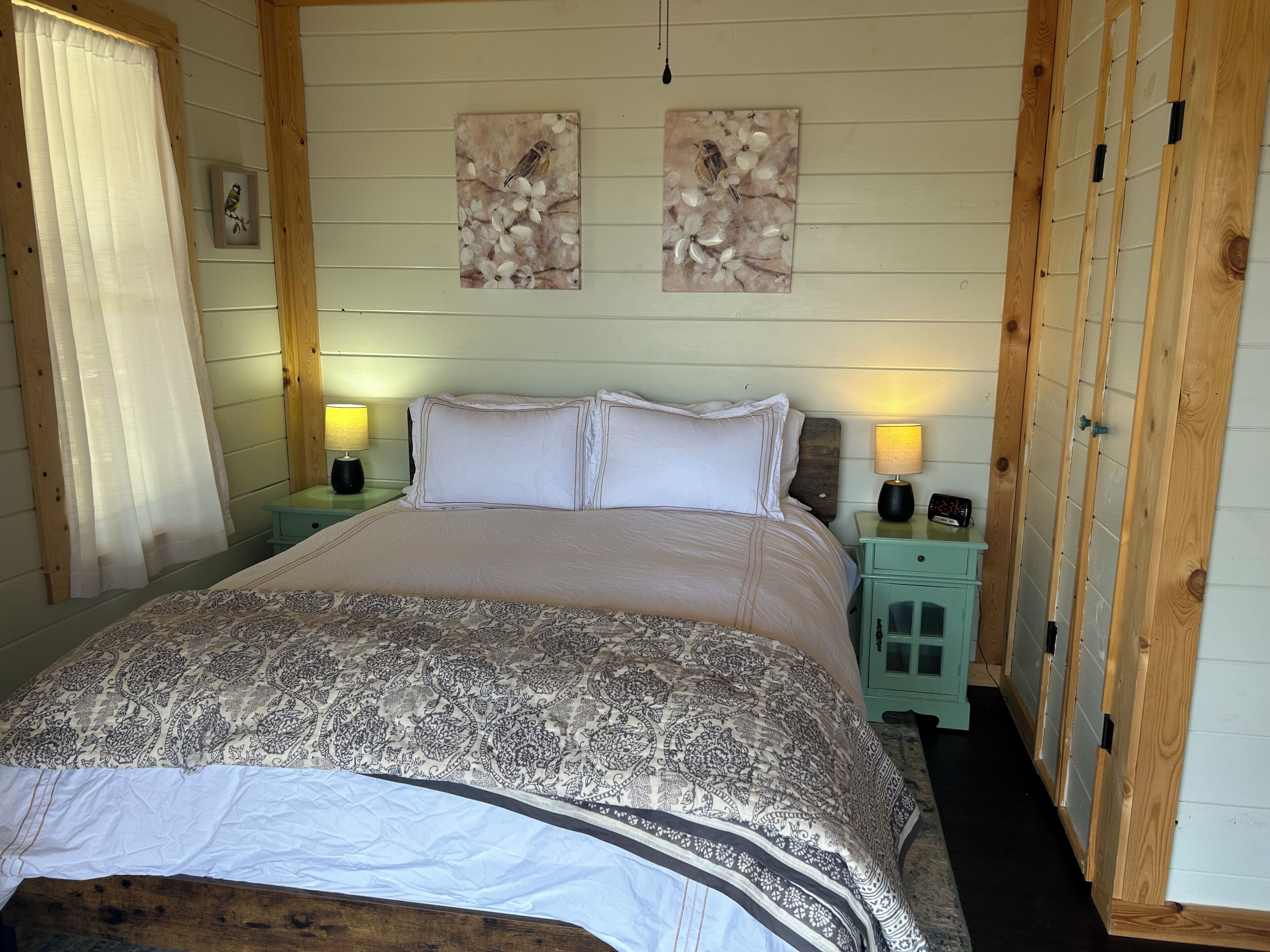 A bedroom with a bed, two nightstands, a window with curtains, and two wall-mounted paintings.