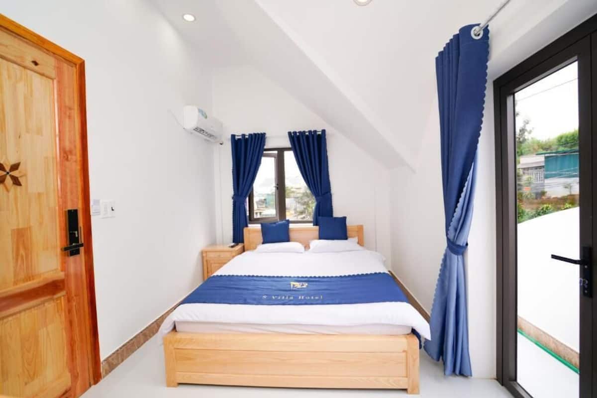 A bedroom with a wooden bed, a window with blue curtains, and a view of the outside.