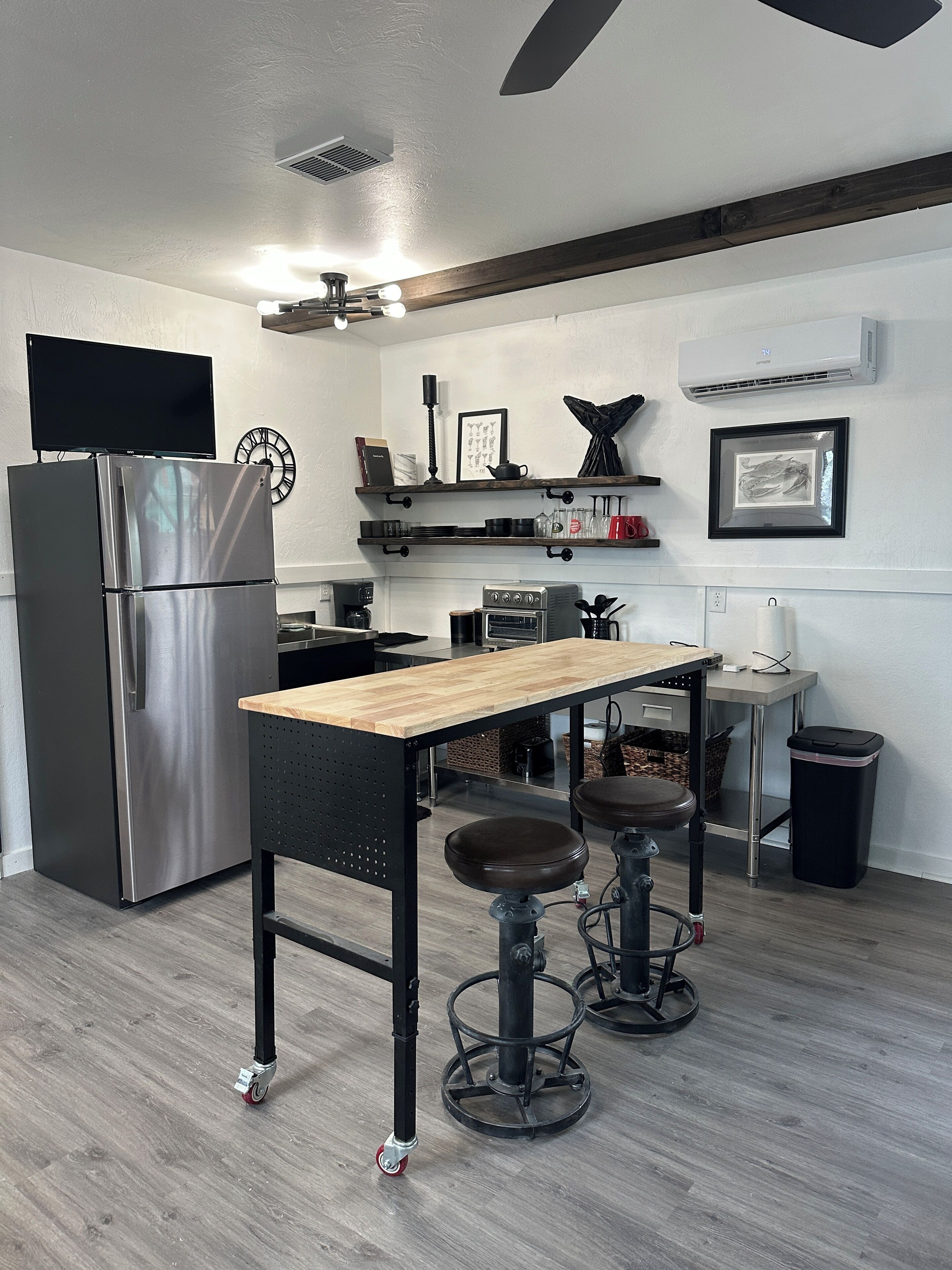 Une cuisine moderne équipée d’un réfrigérateur en acier inoxydable, d’une table en bois, de deux tabourets, d’une cuisinière à gaz et d’un climatiseur fixé au mur.